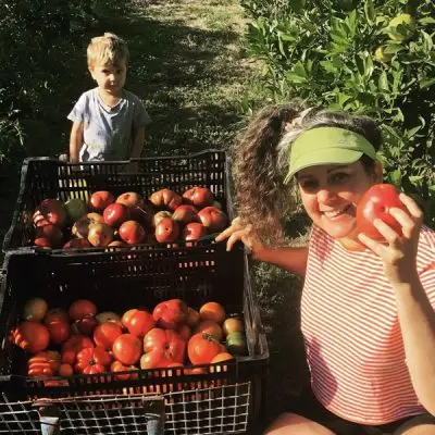 Woman in a green visor shows a red tomato beside crates of apples in a sunlit orchard, with a child standing nearby.
